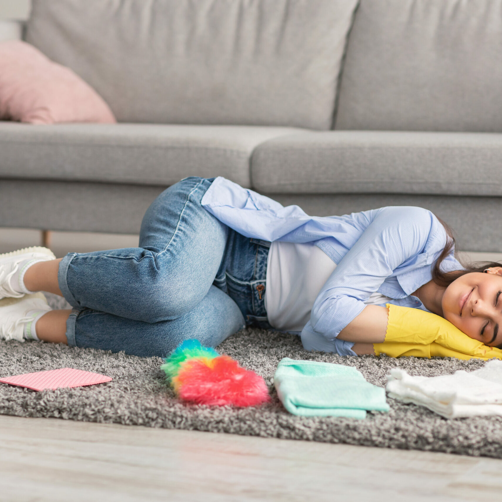 Tired millennial housewife sleeping on gray rug carpet in living room, surrounded by cleaning supplies, copy space. Overworked young lady napping, exhausted after domestic duties, needing rest